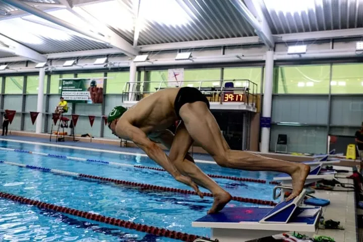 Ryan mid-dive off the blocks, body extended flat over the water at the start of a length.